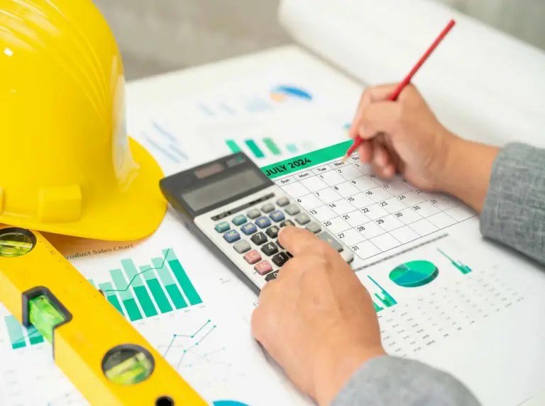 A person uses a calculator and marks dates on a July 2024 calendar surrounded by financial charts and graphs, with a yellow hard hat and spirit level in the foreground, representing construction loan draw schedule planning.