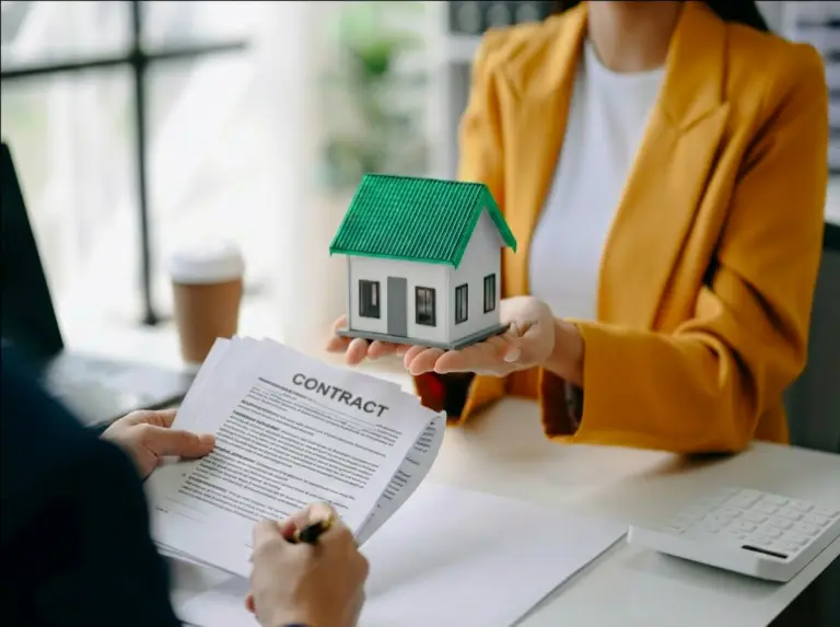 A woman in a yellow blazer holds a small house model while a borrower signs a contract at a desk, illustrating the property-focused approval and closing process involved in securing a hard money loan.